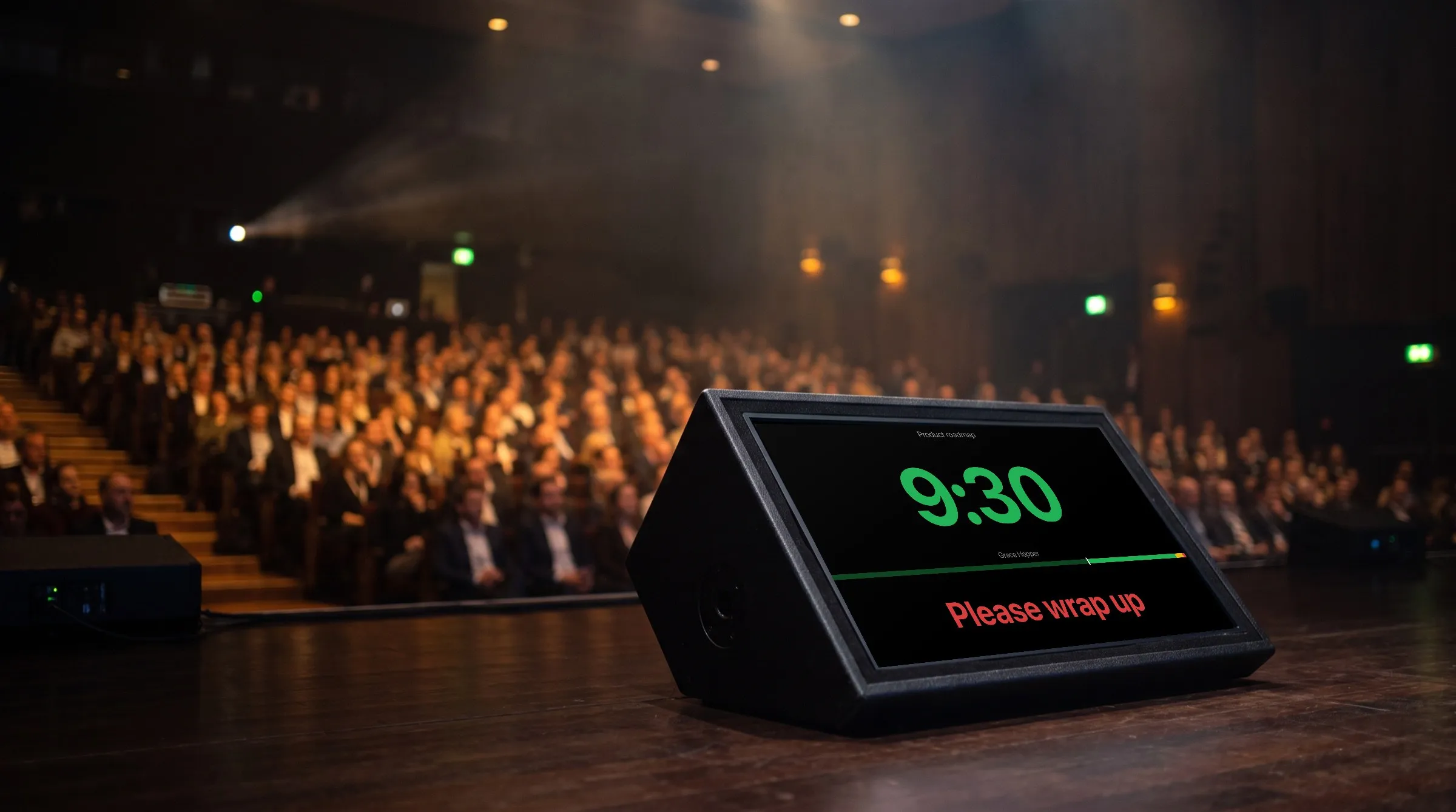 Illustration of a conference stage from the speaker's side: a floor monitor shows CueStride's real screen, 9:30 in green and Please wrap up in red, with the audience out of focus.